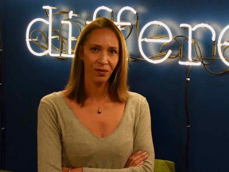 Femme debout devant un mur bleu, bras croisés, portant un haut beige à col en V. Derrière elle, un mot en lettres lumineuses blanches en néon, partiellement lisible, semble être "different".